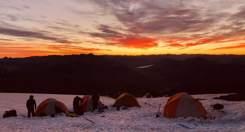 Four tents rest in the snow of a campsite. In the background, the sky appears in vibrant yellow and orange, as the sun rises or sets.
